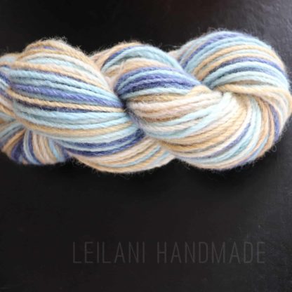 A skein of yarn twisted into a hank, featuring soft pastel shades of blue, cream, beige, and light brown, lies on a black surface. Text reading "LEILANI HANDMADE" appears faintly at the bottom of the image.