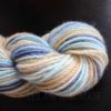 A close-up of a skein of hand-spun yarn with soft, blended colors of light blue, cream, tan, and dark blue. The yarn is twisted and set against a dark background. The texture appears soft and slightly fuzzy.