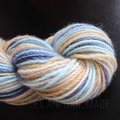 A close-up of a skein of hand-spun yarn with soft, blended colors of light blue, cream, tan, and dark blue. The yarn is twisted and set against a dark background. The texture appears soft and slightly fuzzy.