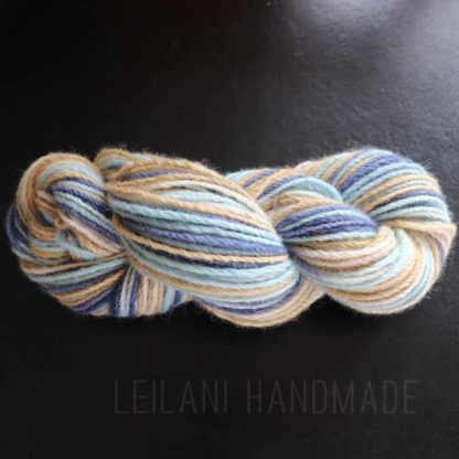 A skein of yarn twisted into a hank, featuring soft, blended shades of cream, beige, light blue, and dark blue, rests on a dark surface. The words "LEILANI HANDMADE" are faintly visible beneath the yarn.