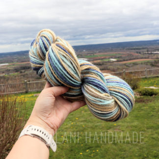 A hand holds a skein of multicolored alpaca handspun yarn with shades of blue, beige, and brown against a scenic outdoor backdrop featuring rolling fields, a wooden fence, and a cloudy sky. The wrist wears a white smartwatch. The text "LEILANI HANDMADE" is at the bottom.