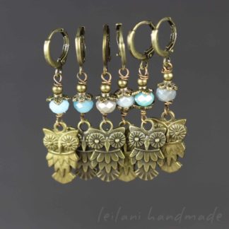 A set of six handmade clip on owl stitch markers featuring brass owl charms. Each pair includes different colored beads: turquoise, grey, and pale blue, above the owl charm. The background is plain and the text "leilani handmade" appears subtly in the lower right corner.