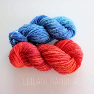 Two skeins of Corriedale handspun hand-dyed yarn are displayed on a white surface. The top skein is a gradient of blue hues, while the bottom skein features shades of red and orange. The text "LEILANI HANDMADE" is faintly visible beneath the skeins.