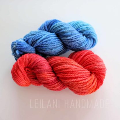Two skeins of Corriedale handspun hand-dyed yarn are displayed on a white surface. The top skein is a gradient of blue hues, while the bottom skein features shades of red and orange. The text "LEILANI HANDMADE" is faintly visible beneath the skeins.