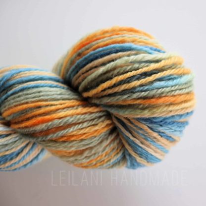 A skein of handspun yarn in soft shades of blue, orange, cream, and green, twisted into a neat coil. The yarn has a smooth, slightly glossy texture, and the colors blend gently, creating a variegated effect against a plain white background.