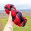 A hand holds a skein of multicolored yarn, predominantly featuring shades of red, pink, black, and beige.