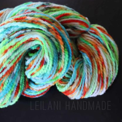 A skein of handspun yarn is coiled on a black surface. The yarn features vibrant colors including green, blue, orange, yellow, and red. Text reading “LEILANI HANDMADE” is faintly visible across the bottom of the image.