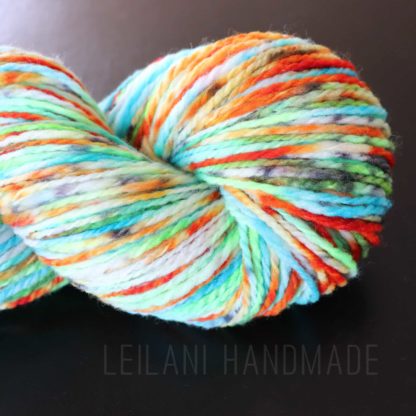 A skein of thick, multicolored yarn with shades of blue, green, orange, yellow, white, and hints of red, displayed against a black background. The yarn has a soft, fluffy texture. The words "LEILANI HANDMADE" appear subtly beneath the skein.