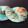 A skein of multicolored yarn with vibrant shades of blue, green, orange, yellow, red, and white is coiled on a black surface. The yarn appears soft and thick. Faint text below the skein reads “LEILANI HANDMADE.”.