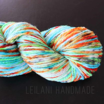 A skein of multicolored yarn with vibrant shades of blue, green, orange, yellow, red, and white is coiled on a black surface. The yarn appears soft and thick. Faint text below the skein reads “LEILANI HANDMADE.”.