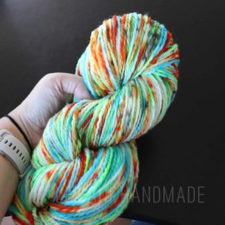 A hand holds a skein of speckle dyed multicolored yarn that features shades of white, green, yellow, orange, and blue. The background is dark, making the vibrant colors of the yarn stand out. The hand is wearing a light gray watch. The words LEILANI HANDMADE is faintly shown at the bottom of the image.