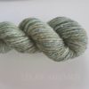 A close-up of a handspun skein of yarn in muted green tones with subtle flecks of yellow, blue, and silver. The yarn is twisted into a neat hank. Faint text at the bottom reads “LEILANI HANDMADE” on a light background.
