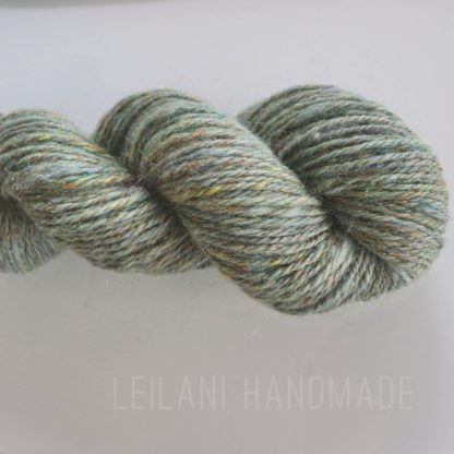 A close-up of a handspun skein of yarn in muted green tones with subtle flecks of yellow, blue, and silver. The yarn is twisted into a neat hank. Faint text at the bottom reads “LEILANI HANDMADE” on a light background.