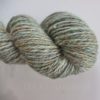 A skein of handspun yarn twisted into a hank, featuring a mix of green, gray, and subtle flecks of blue, yellow, and red. The yarn has a soft, slightly fuzzy texture and a faint metallic shimmer. The background is pale and the text "LEILANI HANDMADE" is faintly visible.