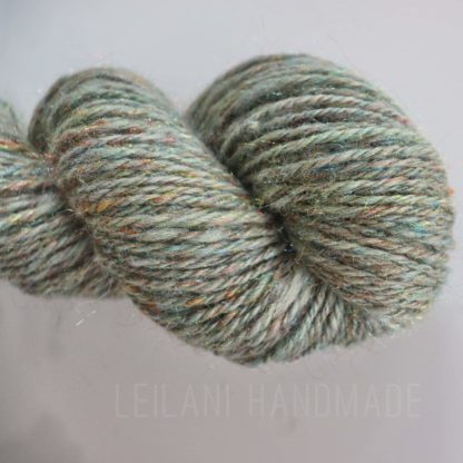 A skein of handspun yarn twisted into a hank, featuring a mix of green, gray, and subtle flecks of blue, yellow, and red. The yarn has a soft, slightly fuzzy texture and a faint metallic shimmer. The background is pale and the text "LEILANI HANDMADE" is faintly visible.