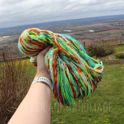 A hand wearing a white watch holds a bundle of multicolored yarn—green, blue, orange, red, and yellow—outdoors. In the background, there is a grassy garden, a fence, and a scenic view of rolling fields under a cloudy sky.