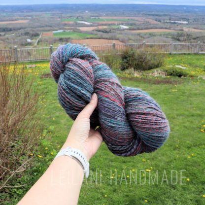 A hand holds a skein of multicolored yarn in front of a scenic rural landscape with green grass, yellow flowers, distant fields, and a wooden fence. The sky is partly cloudy. Text at the bottom reads “LEILANI HANDMADE.”.