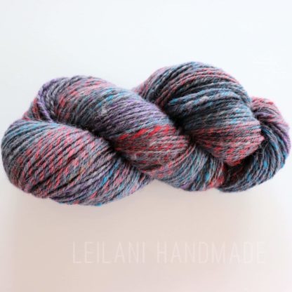 A skein of hand-spun yarn is displayed against a white background. The yarn features a blend of colors, including shades of gray, purple, teal, and red. The texture appears soft and slightly fluffy. The words "LEILANI HANDMADE" are faintly visible below.