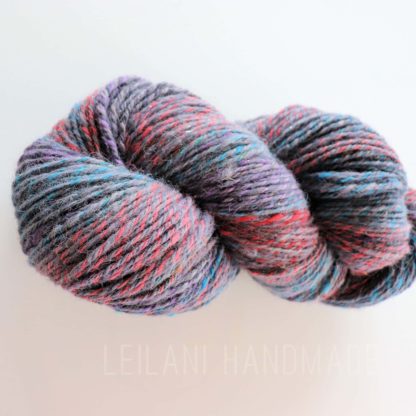 A twisted skein of multicolored yarn featuring shades of gray, blue, purple, and red. The strands are tightly spun together, creating a marled effect. The background is plain white and the words “LEILANI HANDMADE” appear faintly below the yarn.