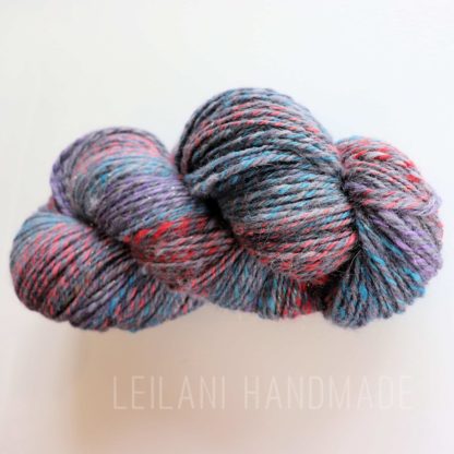 A hank of multicolored, handspun yarn with shades of gray, blue, red, and purple is shown on a white background. The fibers are twisted together, creating a textured, chunky appearance. Faint text at the bottom reads "LEILANI HANDMADE."