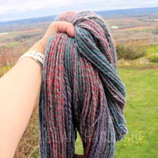 A hand holds up a thick bundle of multicolored yarn with shades of blue, red, gray, and purple. The background shows a grassy field, scattered yellow flowers, a fence, and a distant landscape under a cloudy sky.