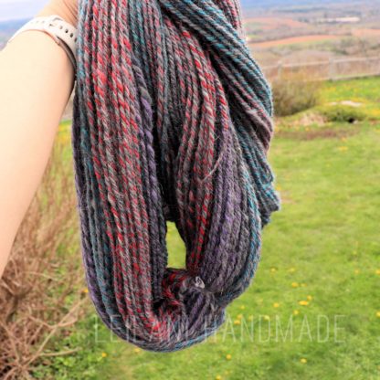 A hand holds a skein of multicolored yarn with twisted strands in red, teal, purple, and gray tones. The background shows a grassy yard with some yellow flowers, blurred trees, and a distant fence under a cloudy sky.