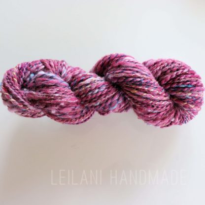 A twisted skein of variegated pink, purple, and blue yarn rests on a white surface. The yarn appears thick and soft, with flecks of white and gold. Light gray text at the bottom reads "LEILANI HANDMADE.