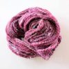 A coiled skein of thick, textured yarn in shades of pink, purple, and white is displayed on a white background. The yarn appears soft and chunky, with visible twists and flecks of various colors throughout. “LEILANI HANDMADE” is faintly visible underneath.
