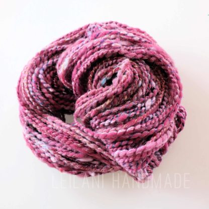 A coiled skein of thick, textured yarn in shades of pink, purple, and white is displayed on a white background. The yarn appears soft and chunky, with visible twists and flecks of various colors throughout. “LEILANI HANDMADE” is faintly visible underneath.