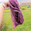 A hand holds up a twisted skein of thick, textured pink and purple yarn outdoors. The background shows a grassy lawn with scattered yellow flowers, a wooden fence, and a blue chair. The words “LEILANI HANDMADE” are faintly visible at the bottom.