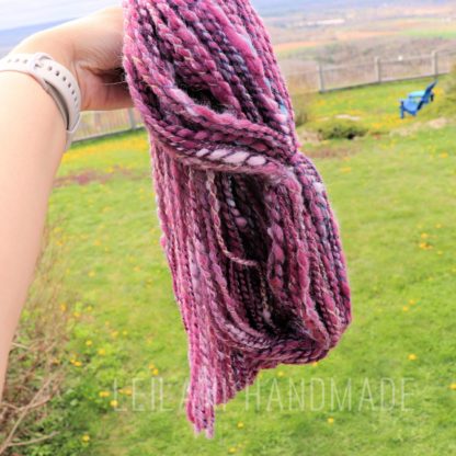 A hand holds up a twisted skein of thick, textured pink and purple yarn outdoors. The background shows a grassy lawn with scattered yellow flowers, a wooden fence, and a blue chair. The words “LEILANI HANDMADE” are faintly visible at the bottom.