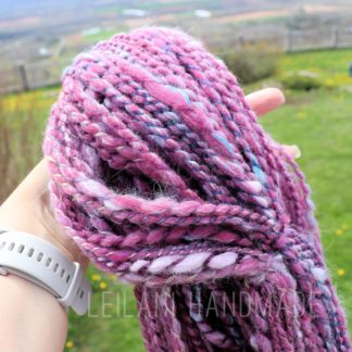 A hand holds a bundle of thick, twisted, pink and purple yarn outdoors. The background is grassy with yellow flowers, bushes, and a distant landscape. The person wears a white smartwatch. "LEILANI HANDMADE" is faintly visible across the image.