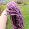 A hand wearing a white watch holds a thick bundle of handspun yarn in shades of pink, purple, black, and white. The background is a green grassy lawn with scattered yellow flowers, a fence, and distant landscape views. “LEILANI HANDMADE” is overlaid.