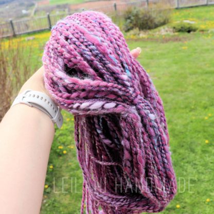 A hand wearing a white watch holds a thick bundle of handspun yarn in shades of pink, purple, black, and white. The background is a green grassy lawn with scattered yellow flowers, a fence, and distant landscape views. “LEILANI HANDMADE” is overlaid.