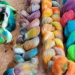 The Ugly Dye experiment: Four bundles of hand-dyed wool roving in various vibrant colors—multicolored, pastel, and rainbow hues—lie on a wooden surface, arranged in both loose and braided forms. The wool appears soft and fluffy.