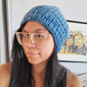 A photo of a tan skinned mixed race woman with long black hair wearing a blue knitted hat and wide white and gold glasses. In the background is a artist painting of a man and woman