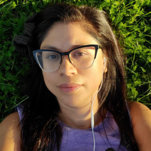 A tanned skinned mixed race woman with long straight black hair and a purple shirt lays in the grass sporting black rimmed glasses and a white headset