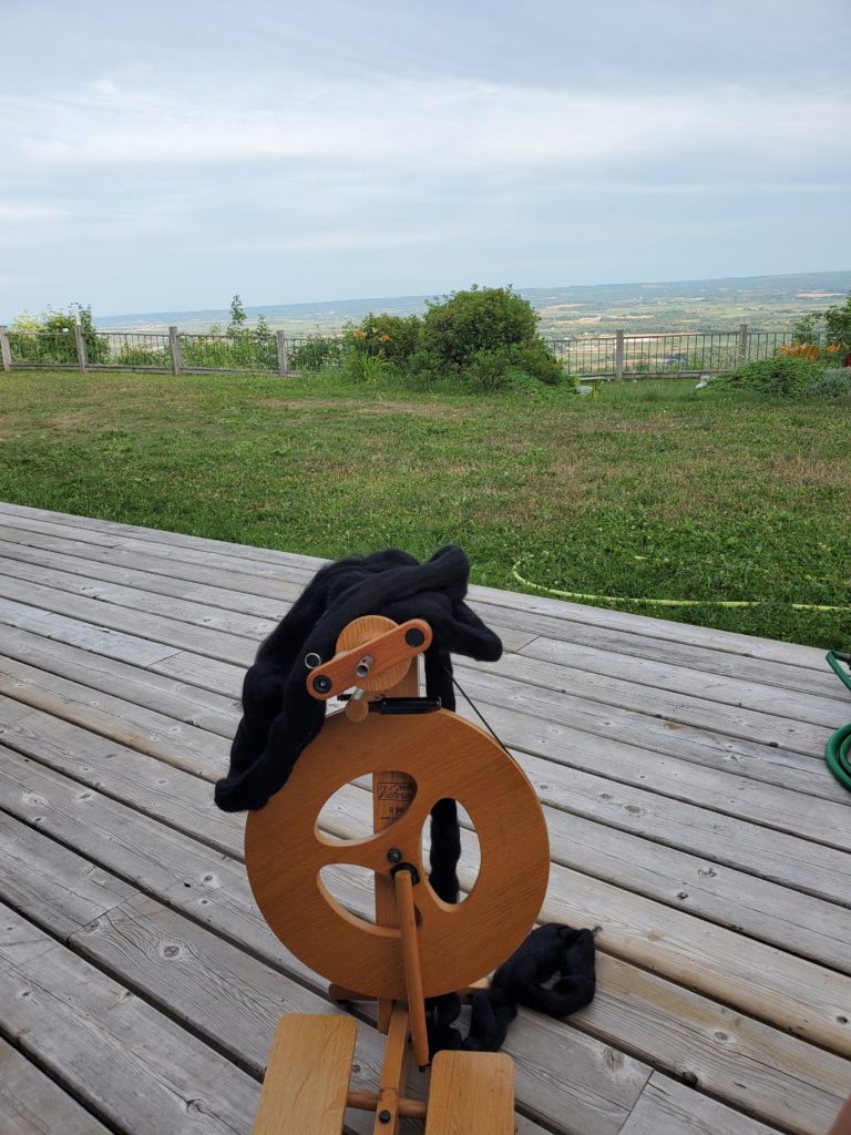 A photo of a small brown foldable spinning wheel with black wool roving on a wooden deck over looking a landscape