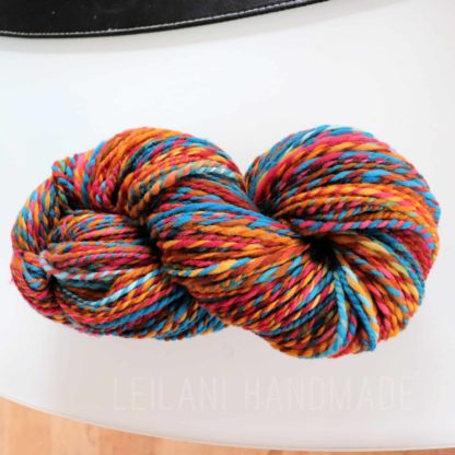 A skein of multicolored yarn with vibrant shades of blue, orange, red, yellow, and pink is coiled on a white surface. The yarn is twisted neatly, showcasing its thick, soft texture. Text on the image reads “LEILANI HANDMADE”.