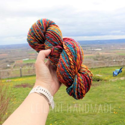A hand wearing a white watch holds a thick, colorful skein of yarn with red, blue, yellow, and green tones. The background features a grassy yard, wooden fence, blue Adirondack chair, and a wide, cloudy sky over distant hills. Text: "LEILANI HANDMADE.
