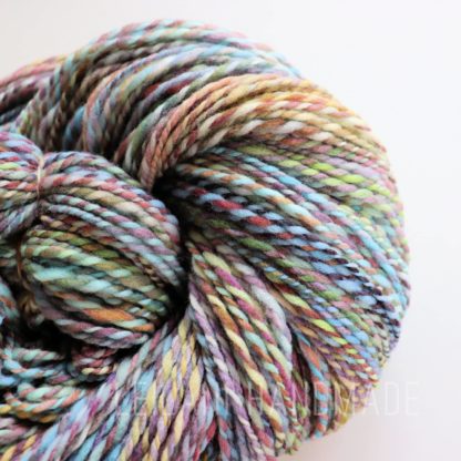 A close-up photo of a Stone hearth handspun yarn. The yarn is twisted and showcases various shades, including blue, green, purple, pink, and yellow. The texture appears soft and plush, highlighting the intricate blending of colors in a spiral pattern. The words LEILANI HANDMADE are visible faintly at the bottom