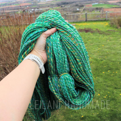 A hand holding a bundle of thick, green variegated yarn in an outdoor setting. The yard has lush green grass and a wooden fence, with a view of distant fields and countryside. The wrist wears a white watch. The text "LEILANI HANDMADE" is faintly visible on the image.