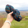 A hand holds up a skein of multicolored yarn against a scenic outdoor backdrop. The yarn features blue, orange, and yellow colors. Text reads "LEILANI HANDMADE" faintly at the bottom