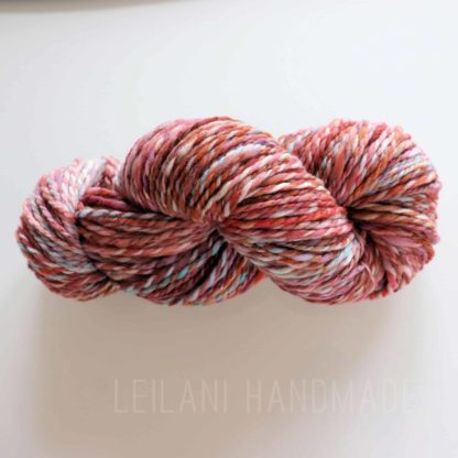 A skein of handspun yarn twisted into a hank, featuring a mix of pink, white, red, and orange tones. The yarn sits on a plain white surface, and the words “LEILANI HANDMADE” appear faintly below it.