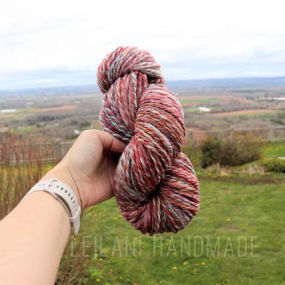 A hand wearing a light gray smartwatch holds a skein of thick, multicolored yarn in front of a grassy field with a scenic, cloudy landscape in the background. Text at the bottom reads “LEILANI HANDMADE.”.