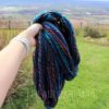A hand wearing a white wristwatch holds a large bundle of colorful, twisted yarn outdoors. The yarn has blue, orange, pink, and purple strands. In the background, there is a grassy yard, a fence, bushes, and a scenic view of fields and distant hills.