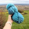 A hand holding a large skein of bright blue yarn outdoors, with a green lawn, some shrubs, and a wide view of rolling fields and a cloudy sky in the background. The words "LEILANI HANDMADE" are faintly overlaid at the bottom of the image.