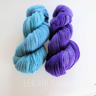 Two neatly twisted skeins of yarn are placed side by side on a white background. The left skein is light blue, and the right skein is bright purple. Both have a soft, smooth texture. Faint text below reads “LEILANI HANDMADE.”.
