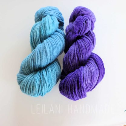 Two neatly twisted skeins of yarn are placed side by side on a white background. The left skein is light blue, and the right skein is bright purple. Both have a soft, smooth texture. Faint text below reads “LEILANI HANDMADE.”.