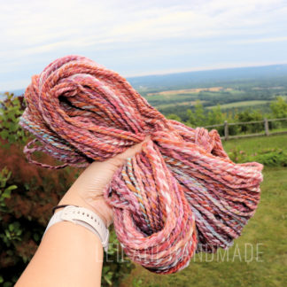 A hand wearing a white watch holds up a thick skein of multicolored, handspun yarn in shades of pink, orange, and purple and blue. The handspun yarn is called "Adventures of Indie Dyeing 2.1." The background shows a green grassy landscape, distant trees, and a cloudy sky, with “LEILANI HANDMADE” faintly visible at the bottom.